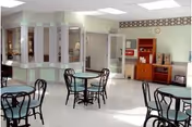 A bright and clean common area with several round tables and black metal chairs. The room has light-colored walls and a white tiled floor. There is a counter area with windows on the left side and a wooden cabinet with a coffee machine and other items on the right side. The ceiling has recessed lighting and skylights.