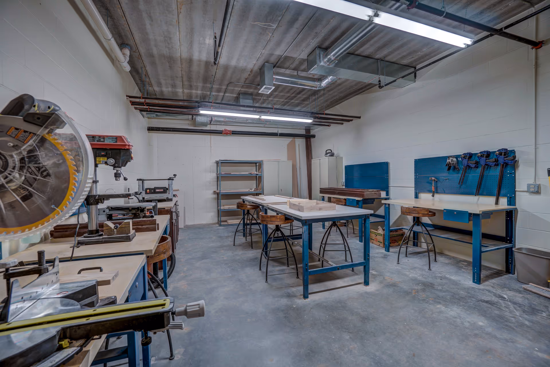 A workshop room with various woodworking tools and equipment including a miter saw, drill press, workbenches, stools, clamps, and storage shelves. The room has concrete floors, white painted walls, and an exposed ceiling with fluorescent lighting.