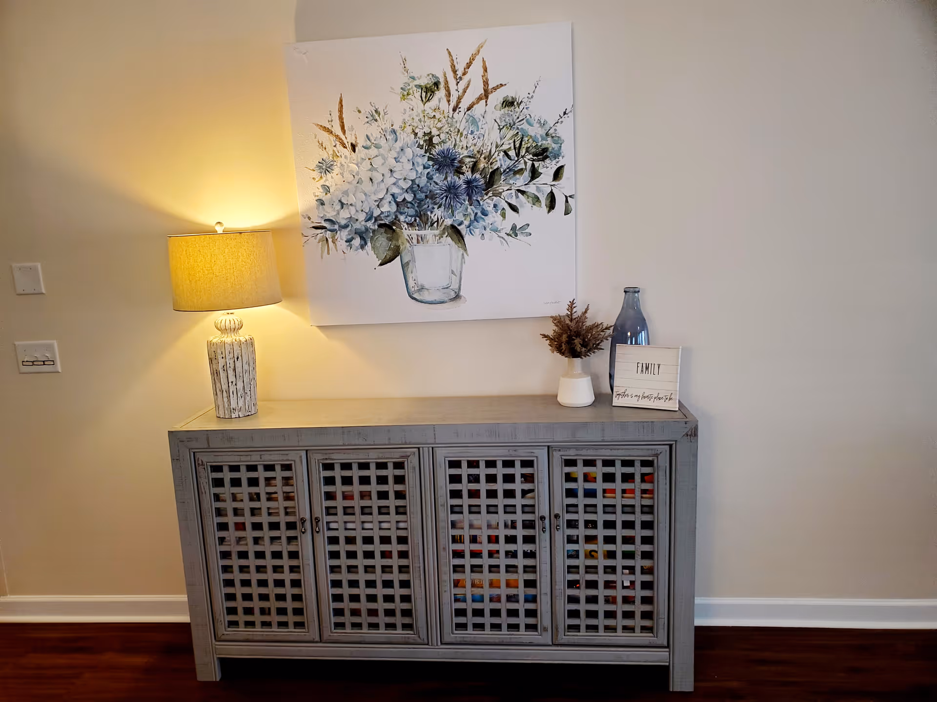 A gray wooden cabinet with lattice-style doors stands against a beige wall. On top of the cabinet is a table lamp with a beige shade, a small white vase with dried flowers, a blue bottle, and a small sign that reads 'FAMILY'. Above the cabinet hangs a painting of a bouquet of blue and white flowers in a glass vase. The floor is dark wood.