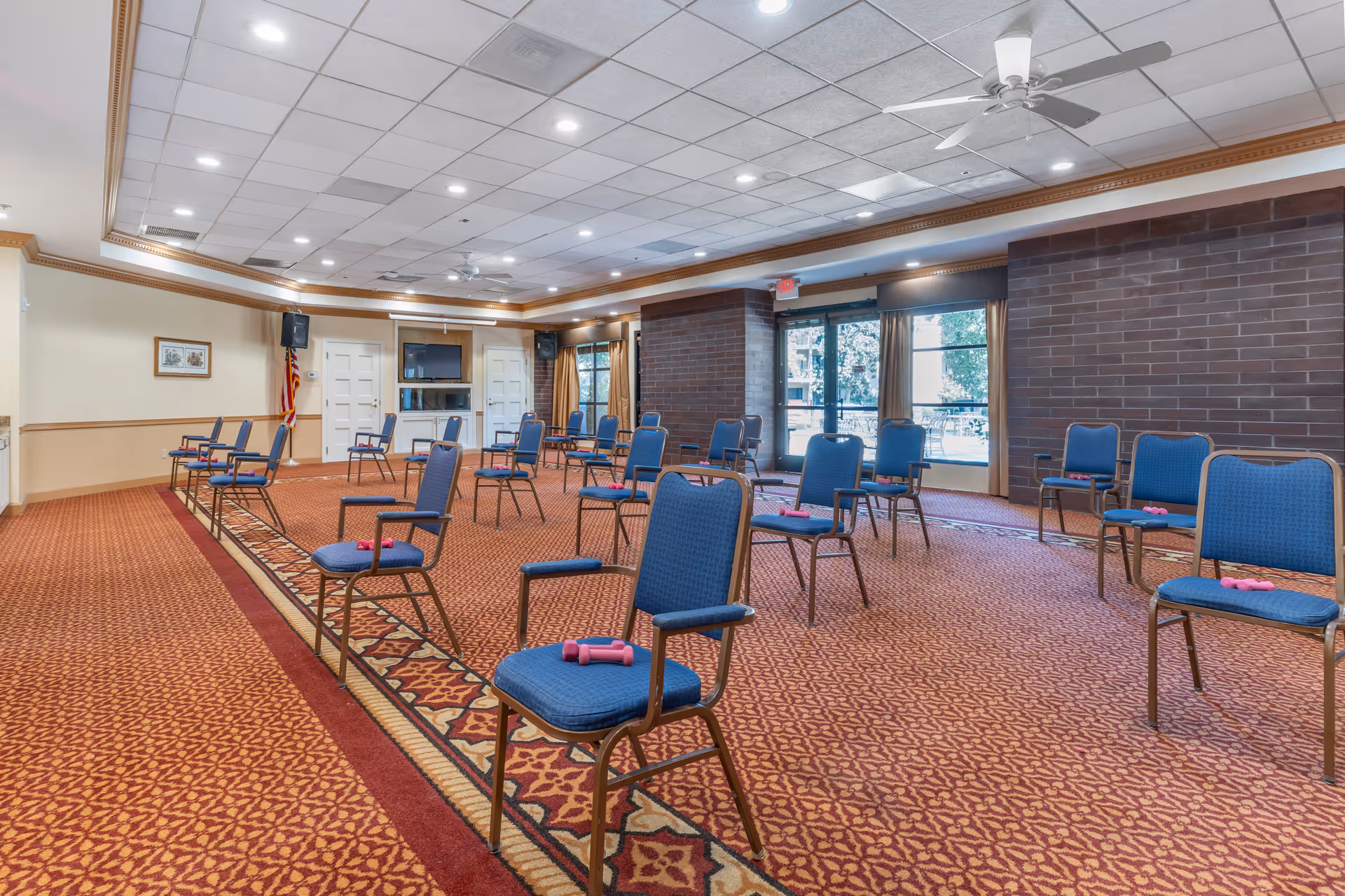 A large room with patterned carpet and blue padded chairs spaced apart, each chair holding a pair of pink hand weights. The room has a drop ceiling with recessed lighting and ceiling fans, a brick accent wall with large windows and glass doors, and an American flag in the corner near a mounted TV and speakers.
