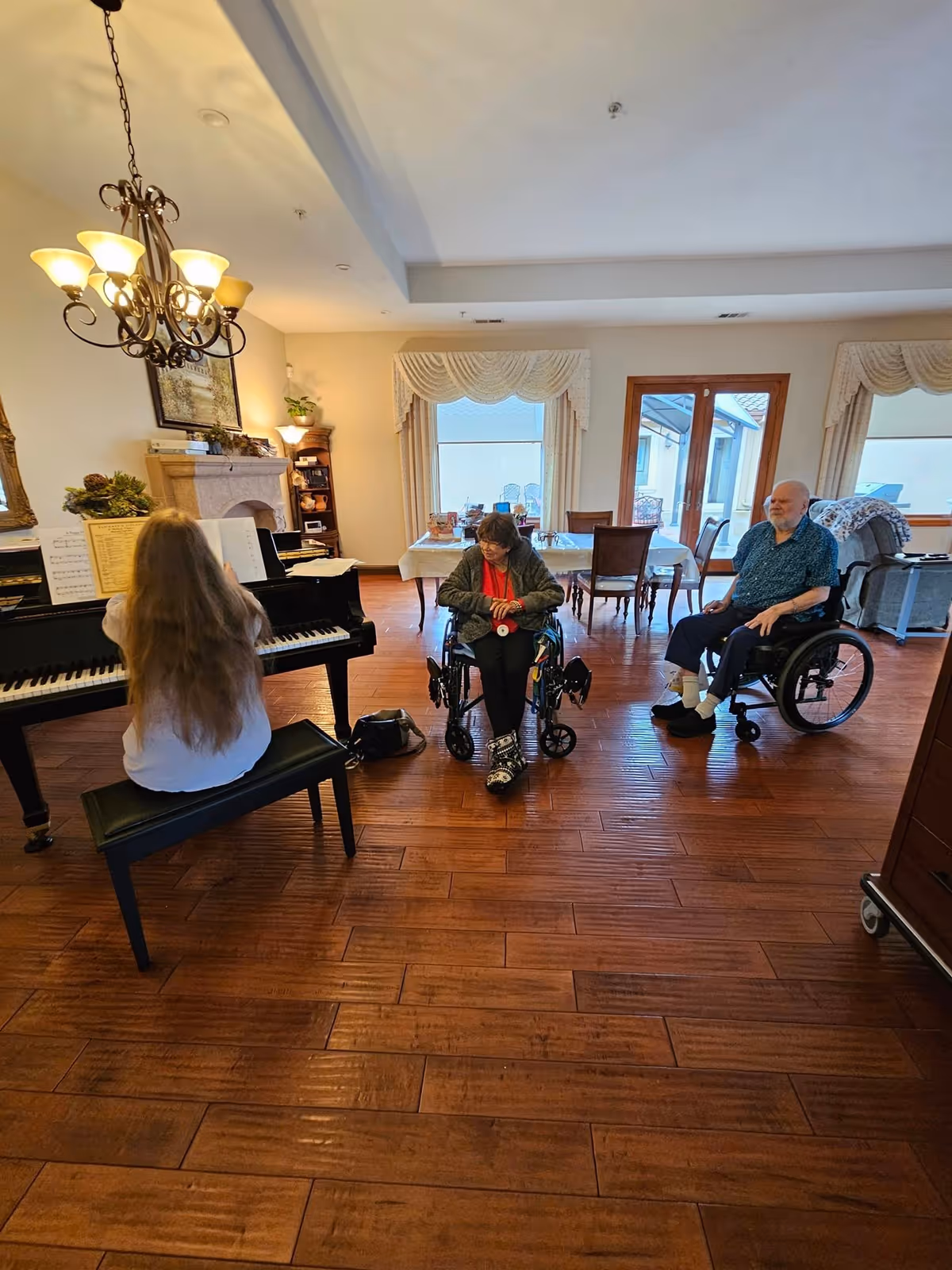 A woman with long hair playing a black grand piano in a room with wooden flooring and a chandelier overhead. Two elderly individuals in wheelchairs are seated facing the piano, listening. The room has large windows with curtains and a dining table with chairs in the background.