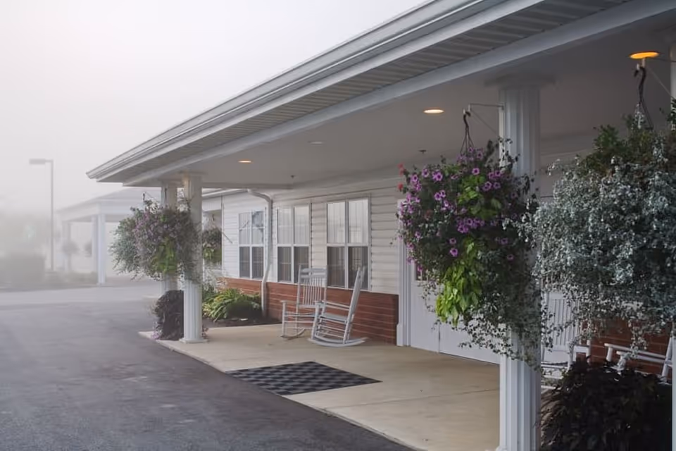 Covered entrance area of a building with white pillars, hanging flower baskets with purple and green plants, two white rocking chairs, and a checkered doormat on the concrete floor. The building exterior has white siding with red brick accents and multiple windows. The scene appears foggy in the background.
