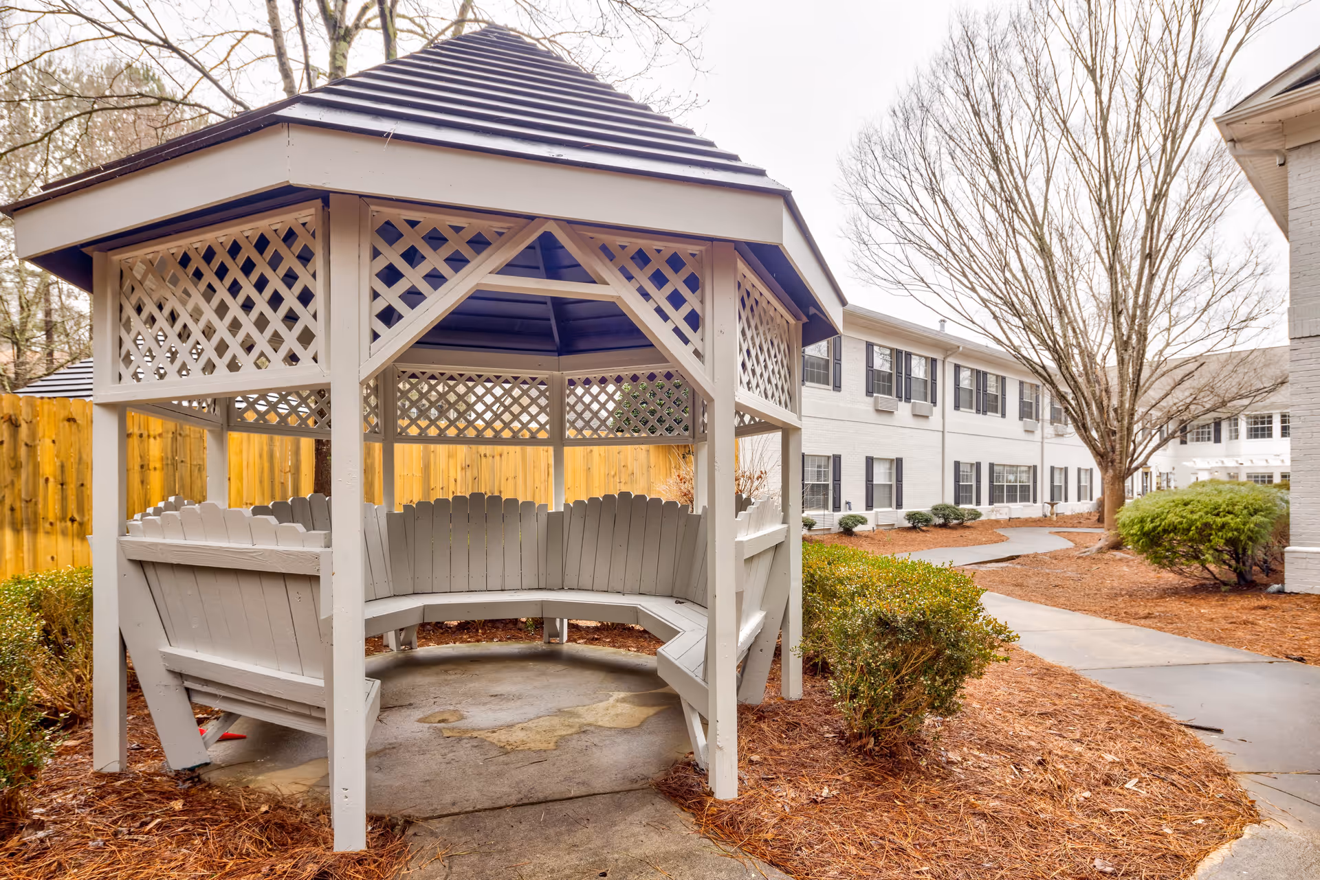 A white wooden gazebo with built-in bench seating is situated outdoors on a concrete pad surrounded by mulch and small bushes. In the background, there is a two-story white building with black window shutters and a paved walkway leading through the landscaped area with leafless trees.