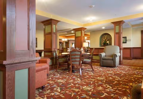 A cozy common area in a senior living facility featuring a patterned carpet with floral designs, wooden columns with green panels, a round wooden table with chairs, an armchair, and soft lighting. The space has warm tones and decorative elements including a niche with flowers on the wall.