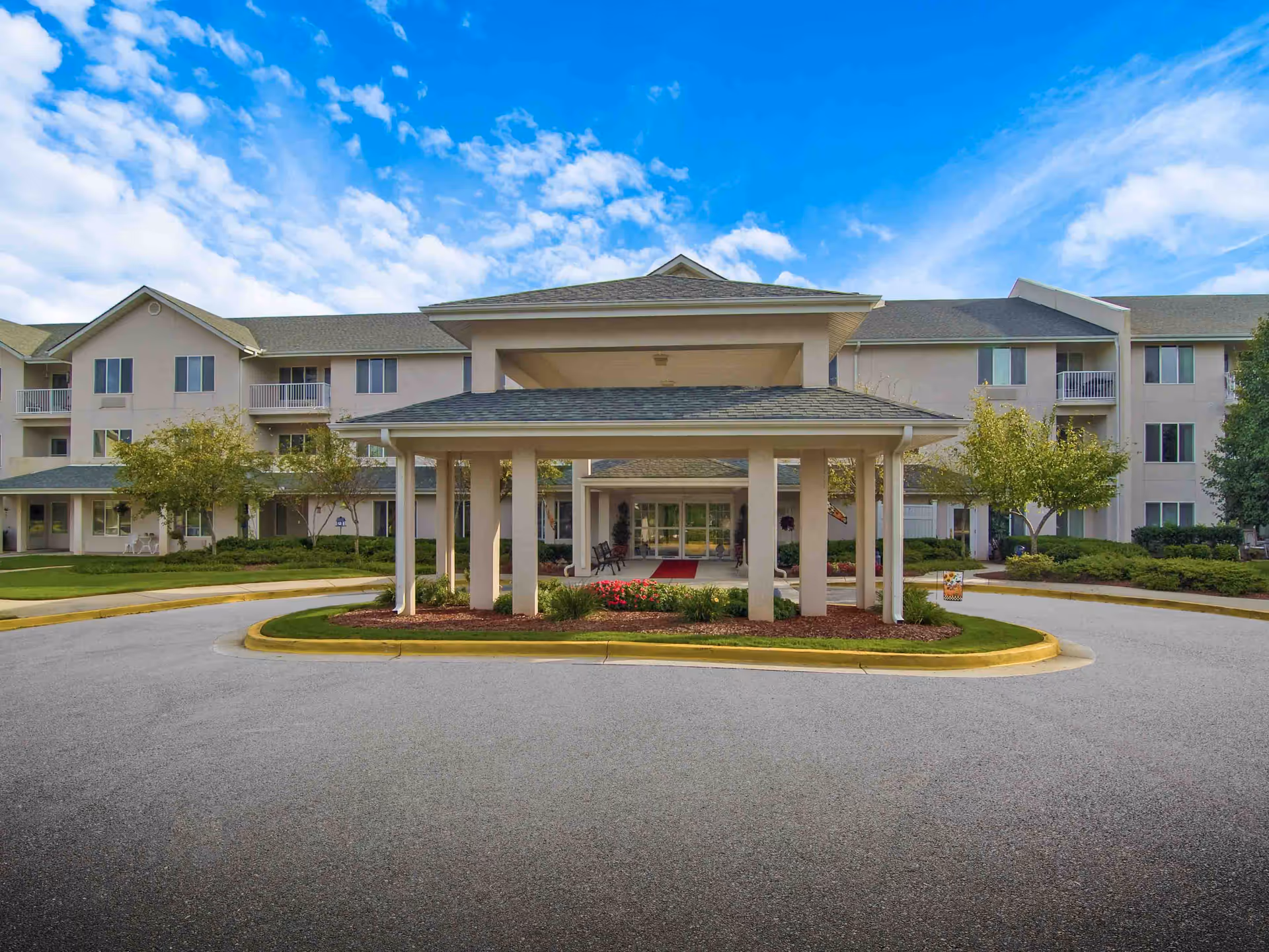 Front entrance of a multi-story senior living building with a covered porte-cochere and landscaped circular driveway under a blue sky.