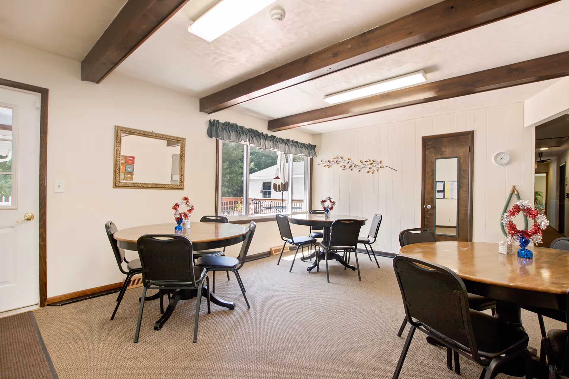 A bright dining room with three round wooden tables, each surrounded by black chairs. Each table has a small blue vase with red and white decorative flowers. The room has beige carpet, white walls with wooden beams on the ceiling, a large window with a green valance, a wall mirror, and a door with a full-length mirror. A clock and a decorative wreath are on the wall near the door.