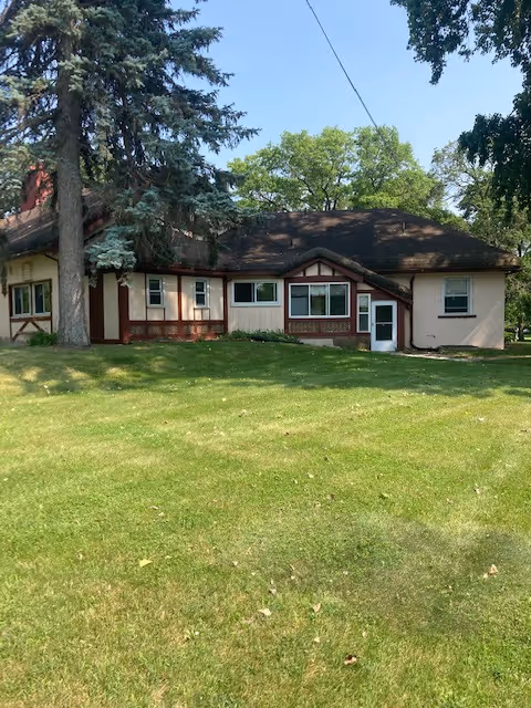 Single-story building with beige walls and brown trim, surrounded by green grass and tall trees under a clear blue sky.