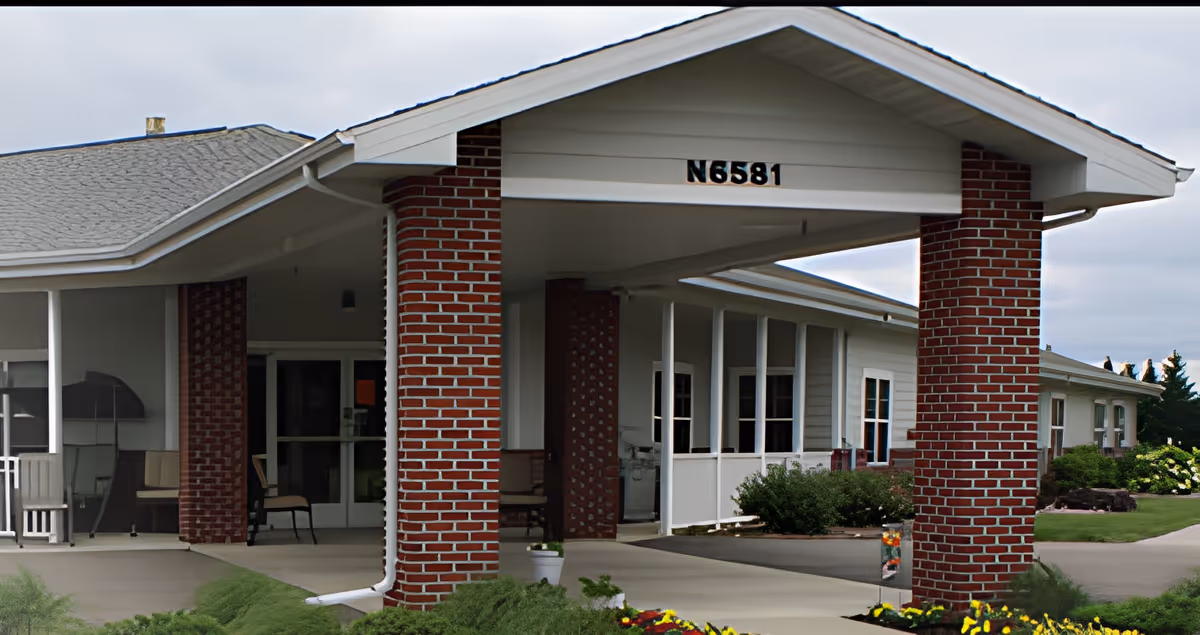 Entrance of a single-story building with a covered porch supported by red brick columns. The building has white siding and multiple windows. There is a paved walkway leading to the entrance, surrounded by green shrubs and a flower bed with yellow and red flowers. The address number N6581 is displayed above the porch.