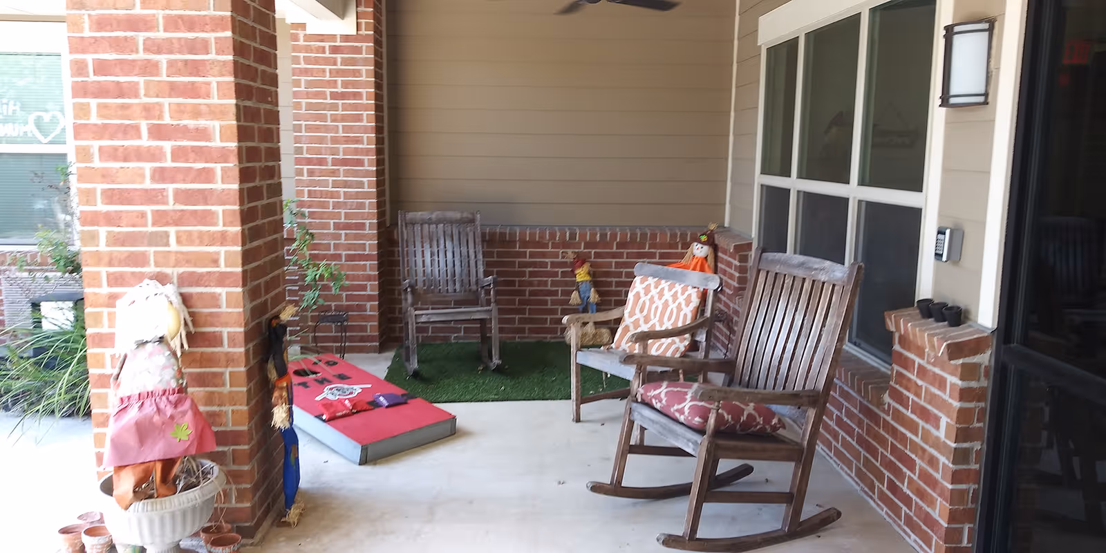 Covered brick porch with wooden rocking chairs, decorative cushions, scarecrow dolls and a cornhole board on the floor.