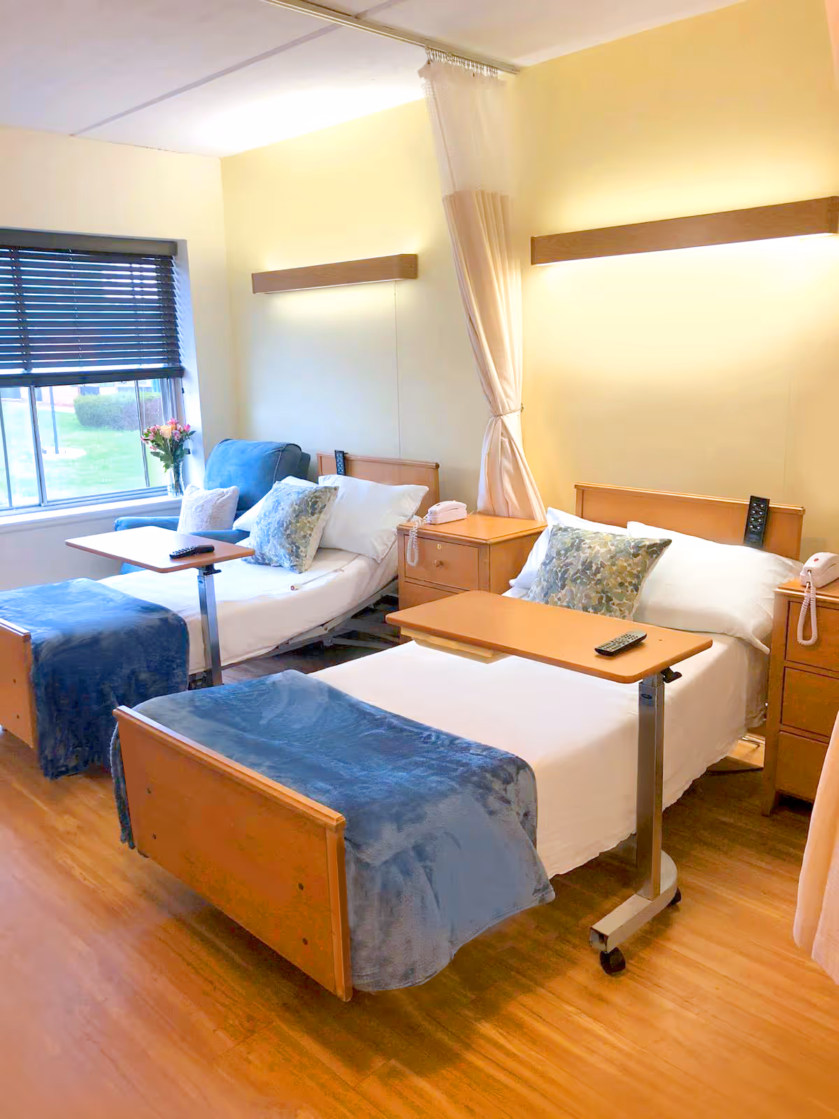 A bright and clean nursing center room with two hospital-style beds, each with a blue blanket and floral pillows. Each bed has an overbed table with a remote control on it. There are wooden nightstands between the beds with telephones on them. A window with blinds and a vase of flowers is visible on the left side, and a privacy curtain is partially drawn between the beds.