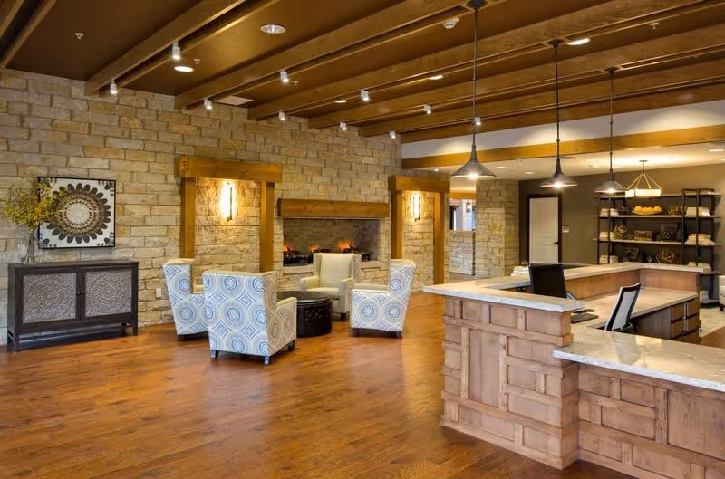 A spacious interior common area with wooden floors and exposed wooden beams on the ceiling. There is a stone wall with a fireplace surrounded by four patterned armchairs and a small round ottoman. To the right, there is a wooden reception desk with marble countertops and two office chairs. Shelving units with decorative items are visible in the background.