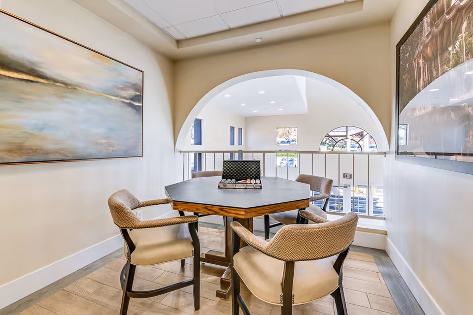 A cozy seating area in a senior living facility featuring a hexagonal table with four cushioned chairs around it. On the table, there is an open case with poker chips. The room has light-colored walls, two large framed paintings, and an arched opening overlooking a bright area with windows and an exit sign.