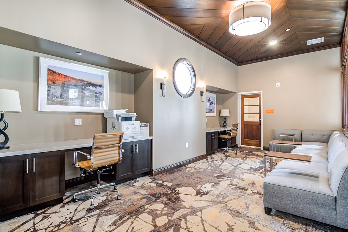 Communal interior lounge with workstations, a printer, leather desk chairs, a long gray sofa, round window and wood-paneled ceiling.