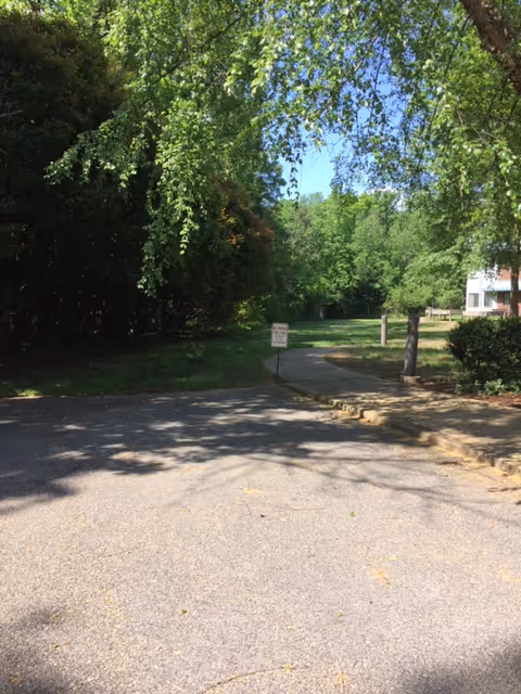 A paved parking area adjacent to a sidewalk leading into a green outdoor space with trees and bushes. A small sign is visible near the sidewalk, and part of a building can be seen on the right side of the image under a clear blue sky.