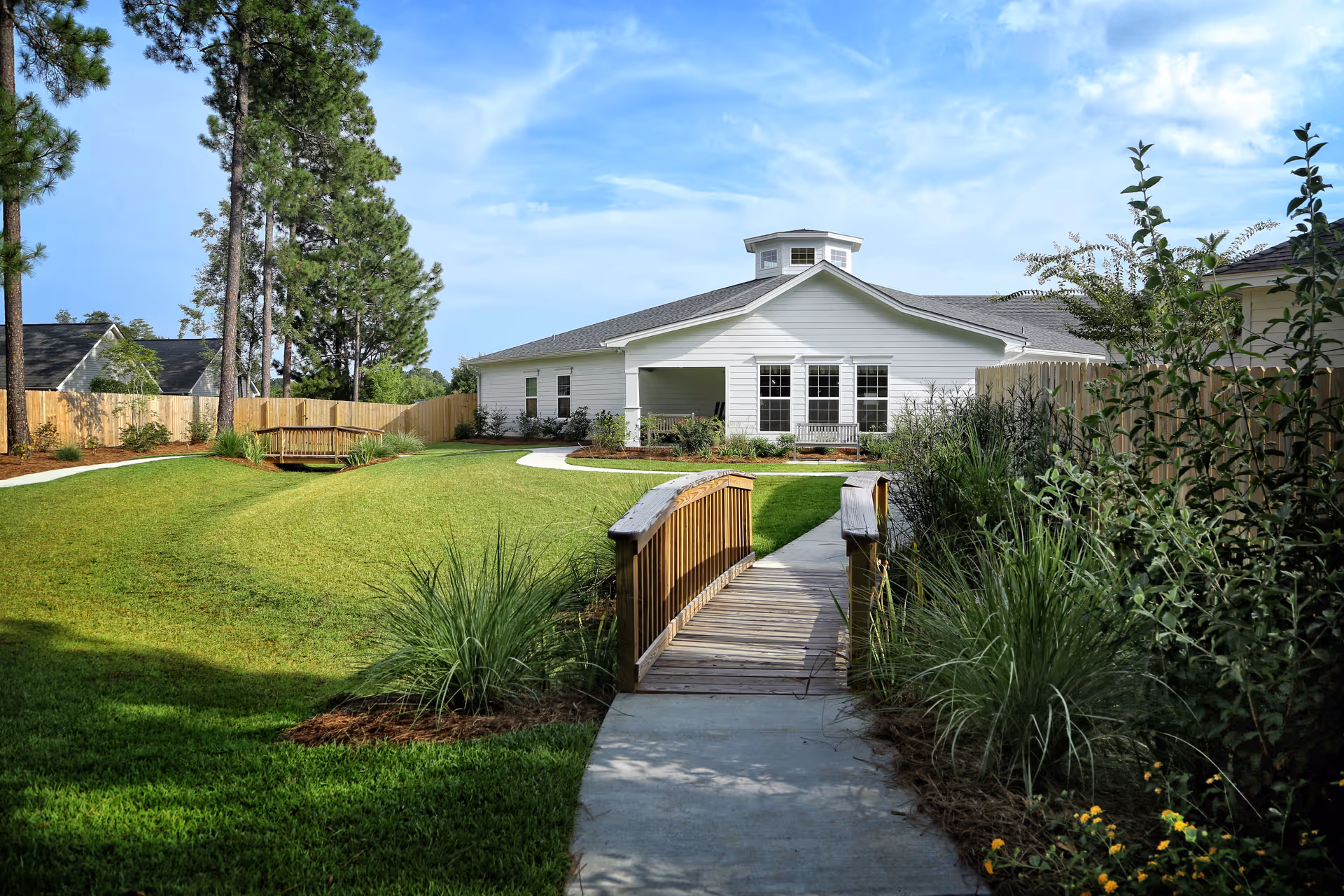 A well-maintained outdoor garden area at Cypress Pond Senior Care featuring a wooden footbridge over a small landscaped area, a paved walkway, green grass, shrubs, and trees surrounding a white building with multiple windows under a blue sky with some clouds.