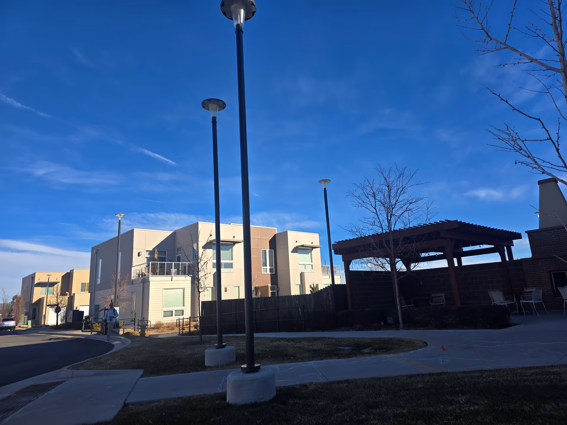 Courtyard of a modern apartment complex with beige multi-story buildings, tall lamp posts, a wooden pergola, and a clear blue sky.