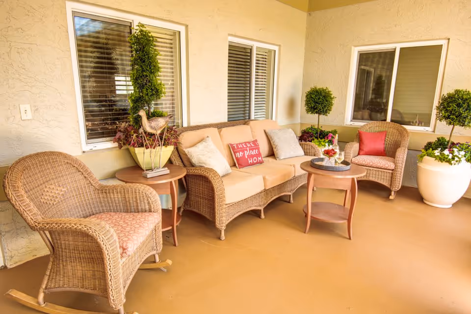 Wicker patio seating with chairs, a sofa, side tables, potted plants, and decorative pillows on a covered porch.
