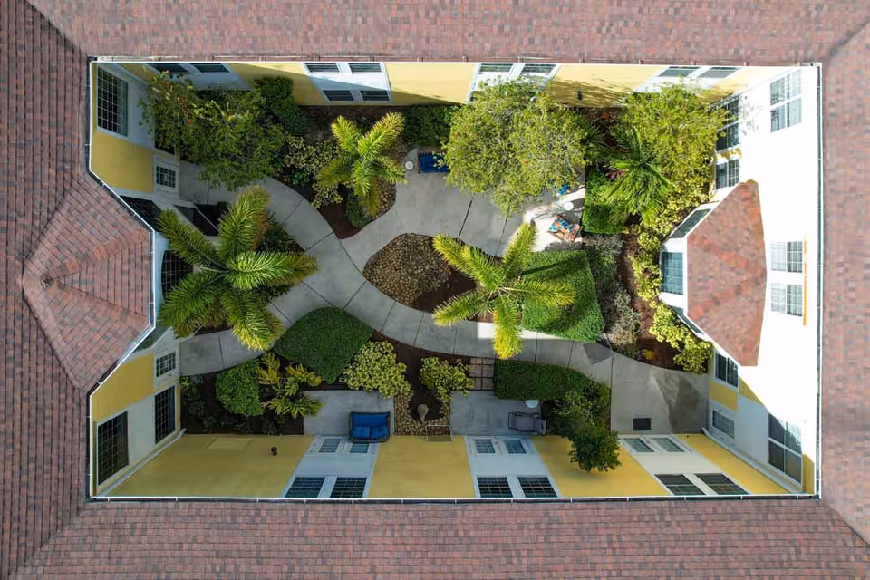 Aerial view of a rectangular courtyard surrounded by a building with yellow walls and red-tiled roof. The courtyard features paved walkways, green bushes, palm trees, and some outdoor seating areas.