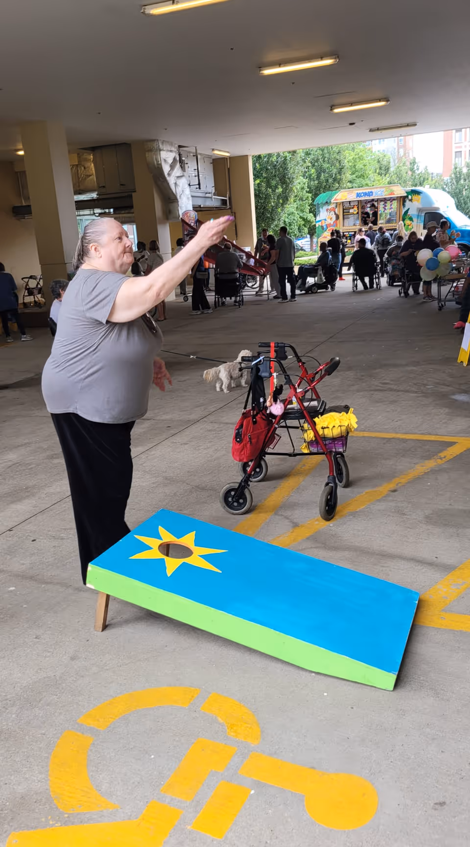 An elderly woman playing a bean bag toss game in a covered outdoor area. She is standing next to a red walker with a basket containing yellow items. In the background, there are several people sitting and standing near an ice cream truck and balloons, with trees and buildings visible outside.
