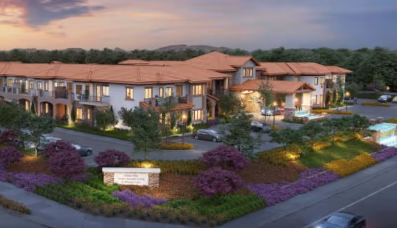 Exterior view of a senior living facility named Varenita of Temecula during dusk, showing a large two-story building with a terracotta roof, surrounded by landscaped gardens with purple and green plants, trees, and a parking area with several cars.