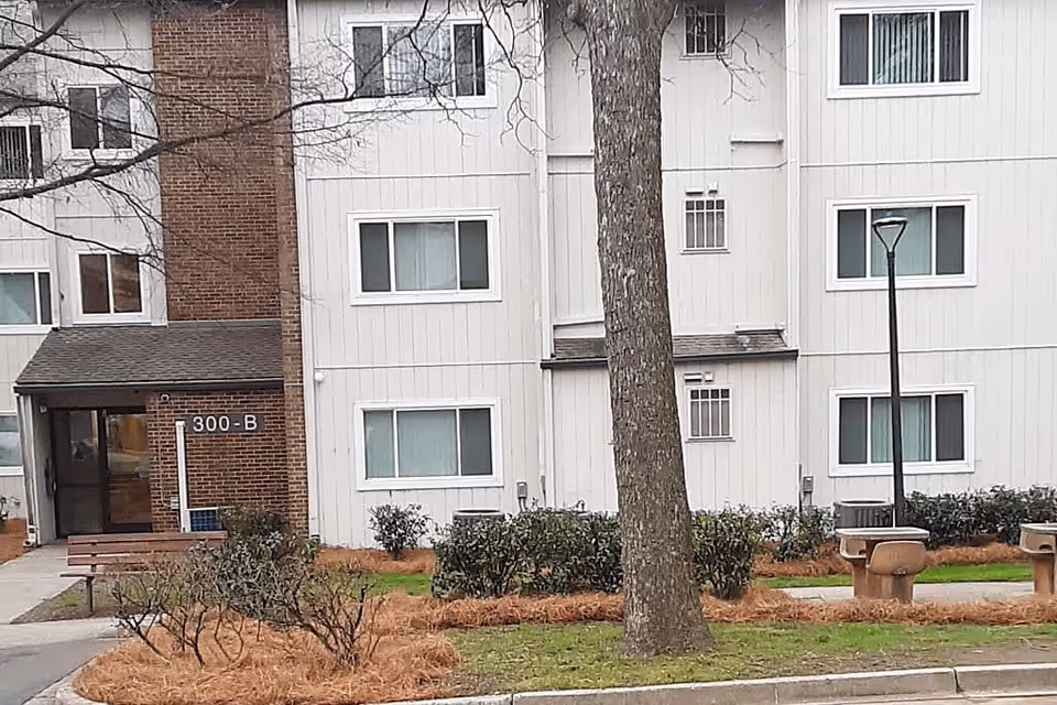 Exterior view of a multi-story residential building with white siding and brick accents. The building has several windows and a main entrance labeled 300-B. In front of the building, there is a landscaped area with bushes, a tree, a street lamp, and two concrete benches.
