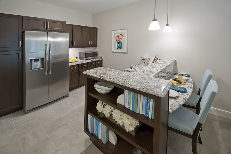Modern kitchen area with dark wood cabinets, a stainless steel refrigerator, a granite countertop with an overhang for seating, two light blue chairs, a small sink, and a built-in bookshelf underneath the counter holding books and decorative items. A framed floral picture hangs on the wall.