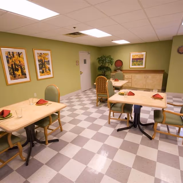 Small dining room with four set tables, green upholstered chairs, checkered floor, framed artwork and a sideboard against a green wall.