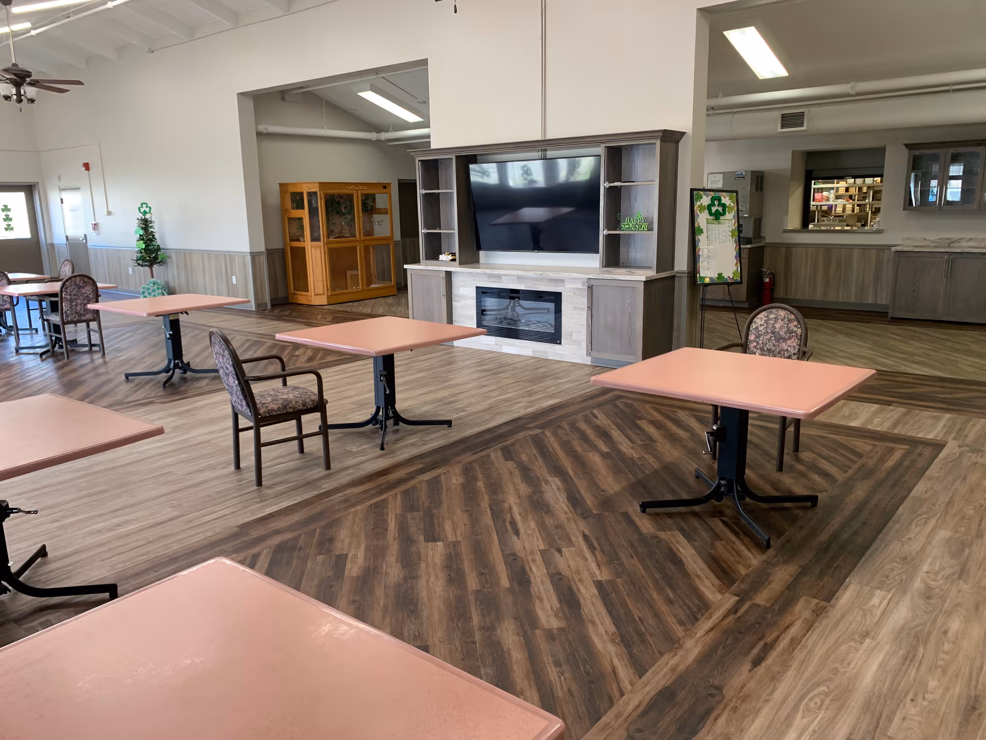 A spacious senior living facility common area with several pink square tables and floral cushioned chairs arranged on a wood-patterned floor. In the background, there is a large entertainment center with a flat-screen TV and an electric fireplace. The room has high ceilings with exposed beams, ceiling fans, and a bulletin board with a shamrock decoration.