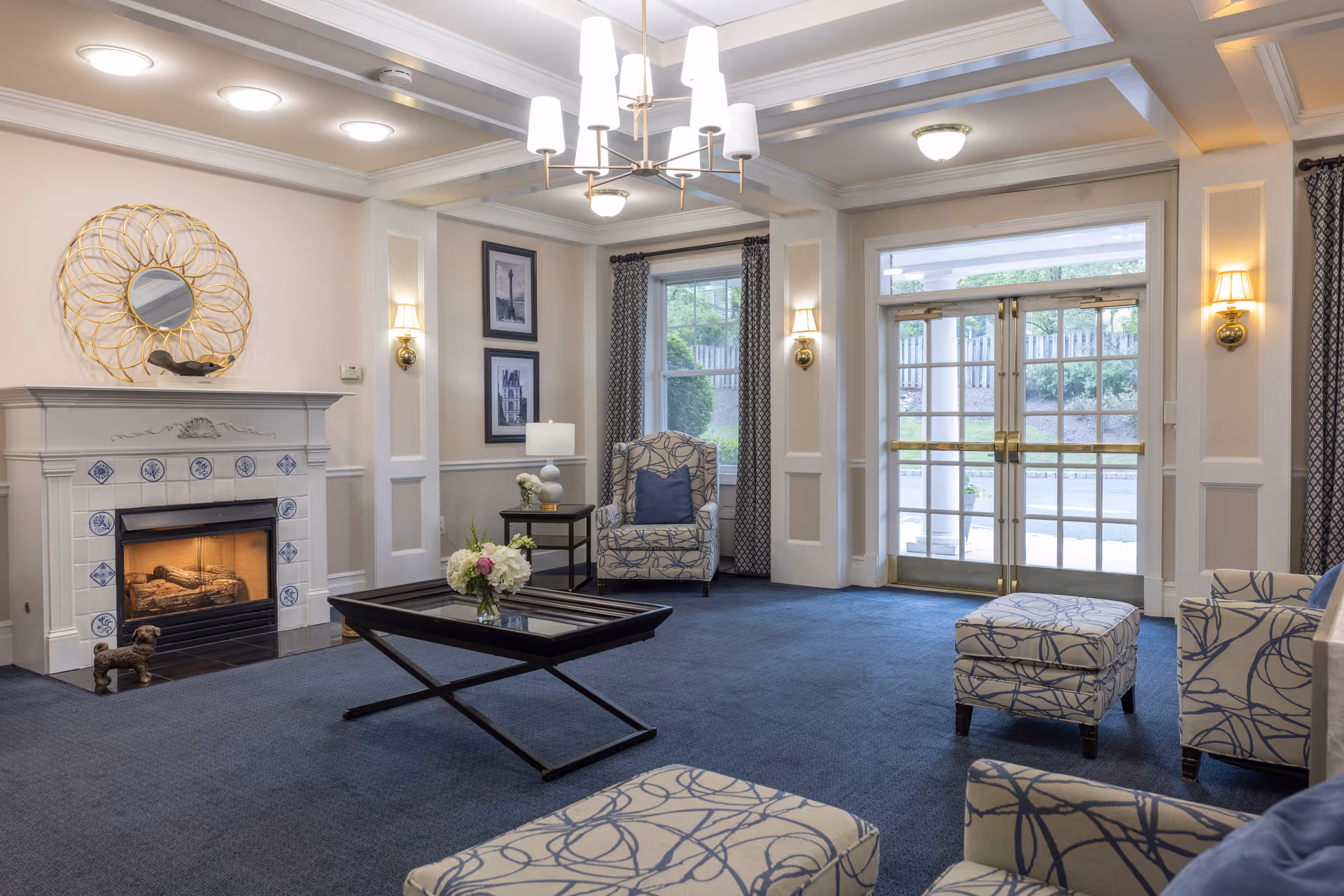A cozy and elegant living room area in an assisted living facility featuring a fireplace with decorative tiles, a round gold-framed mirror above it, patterned armchairs with matching ottomans, a glass coffee table with a flower vase, wall sconces, framed artwork, large windows with curtains, and double glass doors leading outside.
