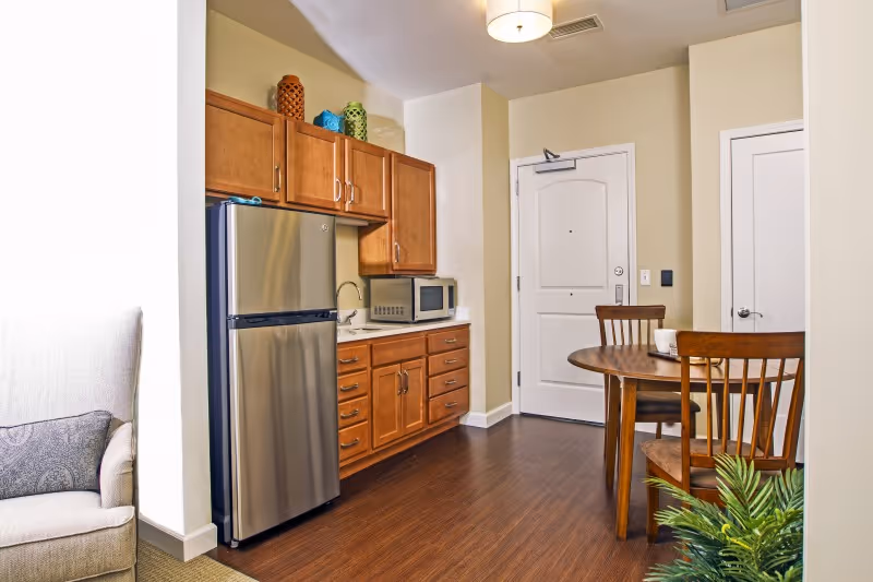 A small kitchen area with wooden cabinets, a stainless steel refrigerator, a microwave on the counter, and a sink. To the right, there is a round wooden dining table with two wooden chairs. The entrance door is visible in the background, and part of a beige armchair with a patterned pillow is seen on the left side. A green plant is in the bottom right corner.