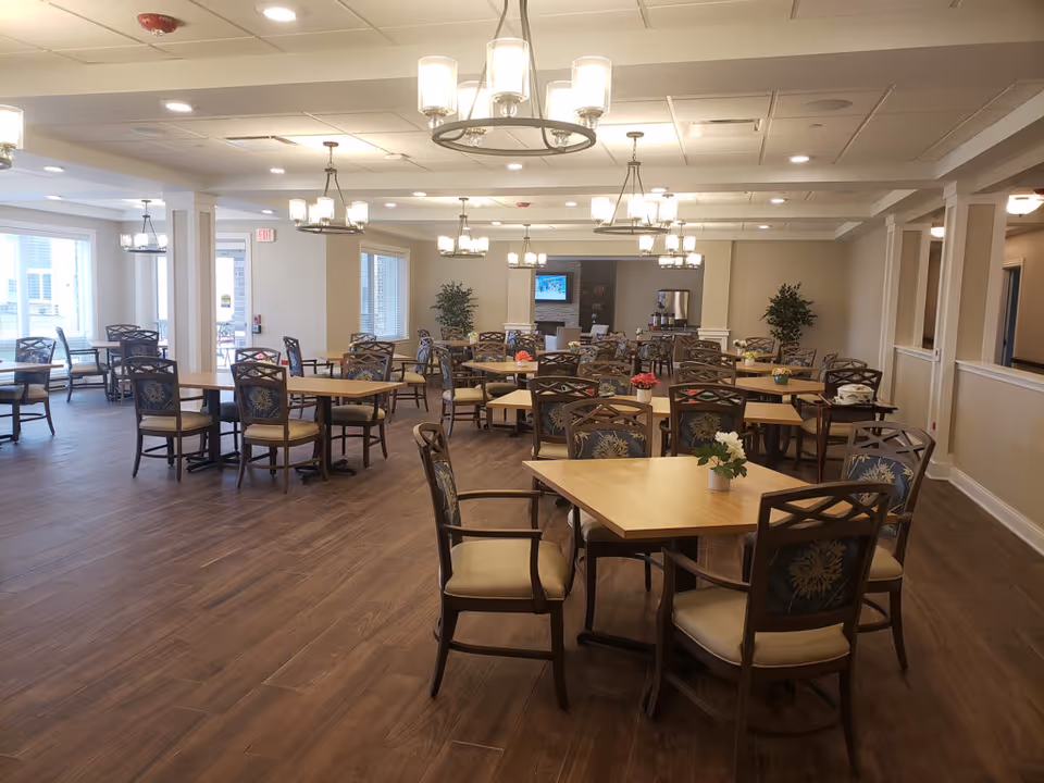A spacious dining room in a senior living facility with multiple wooden tables and cushioned chairs arranged neatly. The room has large windows allowing natural light, several ceiling light fixtures, and decorative plants. A television and a coffee station are visible in the background.