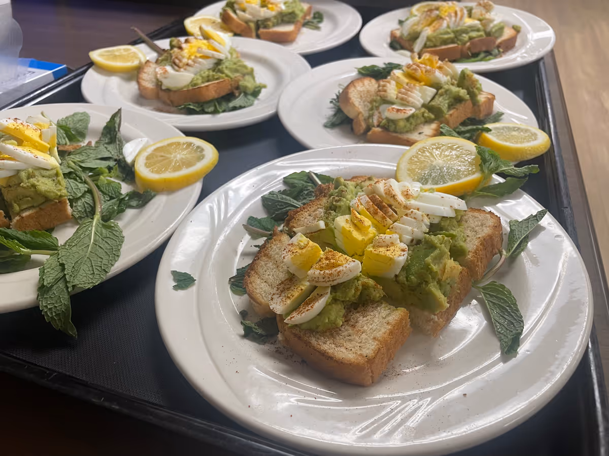 Multiple plates of avocado toast topped with sliced hard-boiled egg, garnished with mint leaves and lemon wedges on a serving tray.