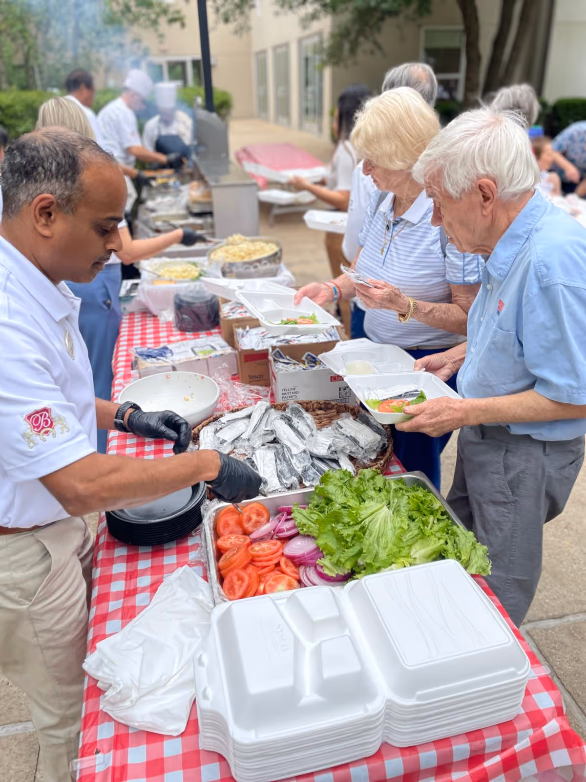 An outdoor buffet setup with a man serving food to elderly people holding white disposable plates. The table is covered with a red and white checkered tablecloth and has trays of sliced tomatoes, onions, lettuce, and wrapped food items. Several people are lined up to receive food, and chefs in white uniforms are visible in the background preparing food.
