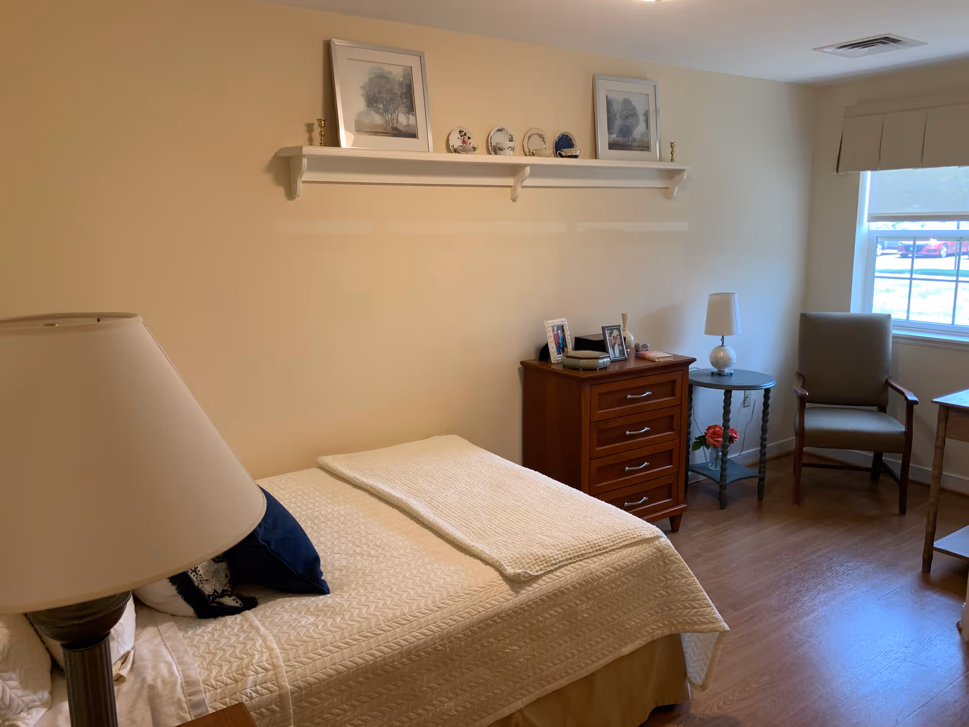 A cozy bedroom with a neatly made bed covered with a cream quilt and a navy blue pillow. A wooden nightstand with a lamp is beside the bed. On the opposite wall, there is a wooden chest of drawers with framed photos and decorative items on top. Above the chest is a white wall shelf holding framed pictures and decorative plates. A small round table with a lamp and a flower vase is next to a cushioned armchair near a window with a view of parked cars outside. The room has wooden flooring and beige walls.