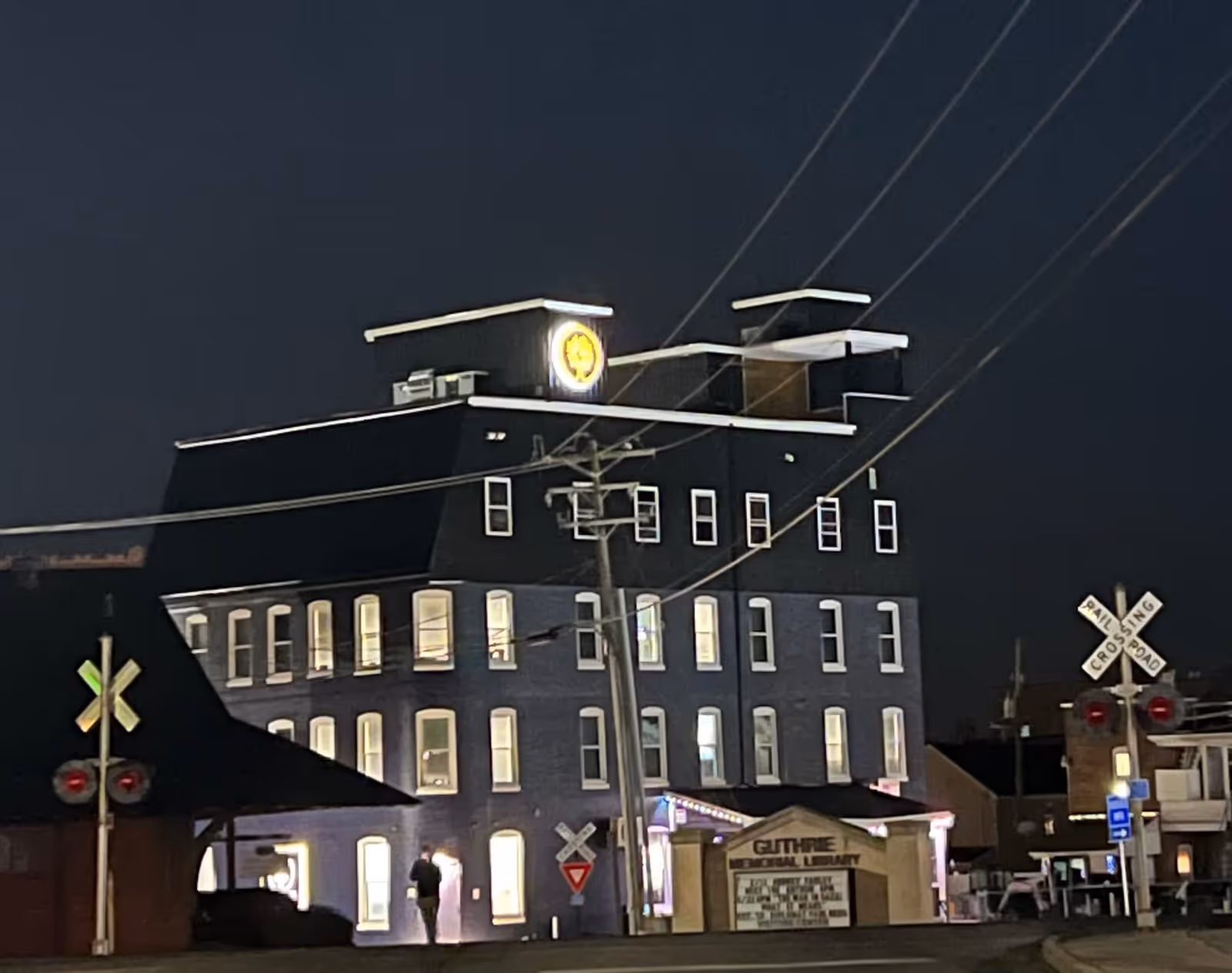 Nighttime view of a multi-story building with many lit windows and a clock on the top. In front of the building is a sign that reads 'Guthrie Memorial Library' and a railroad crossing sign with red lights. Power lines and a utility pole are visible in the foreground.