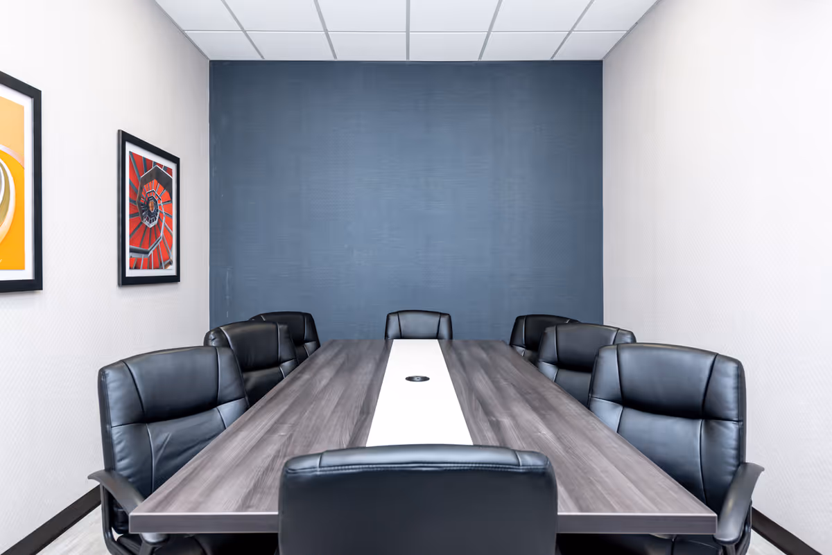 A modern conference room with a long rectangular wooden table surrounded by eight black leather office chairs. The room has a blue accent wall at the far end and two framed abstract artworks on the left wall. The ceiling has white tiles with recessed lighting.