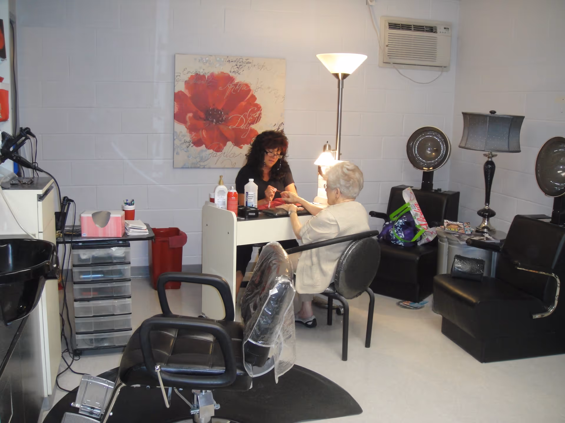 A salon room with a woman providing a manicure to an elderly woman seated at a small table. The room has salon chairs, hair dryers, a floor lamp, and a large red flower painting on the wall. There is an air conditioning unit mounted on the wall.
