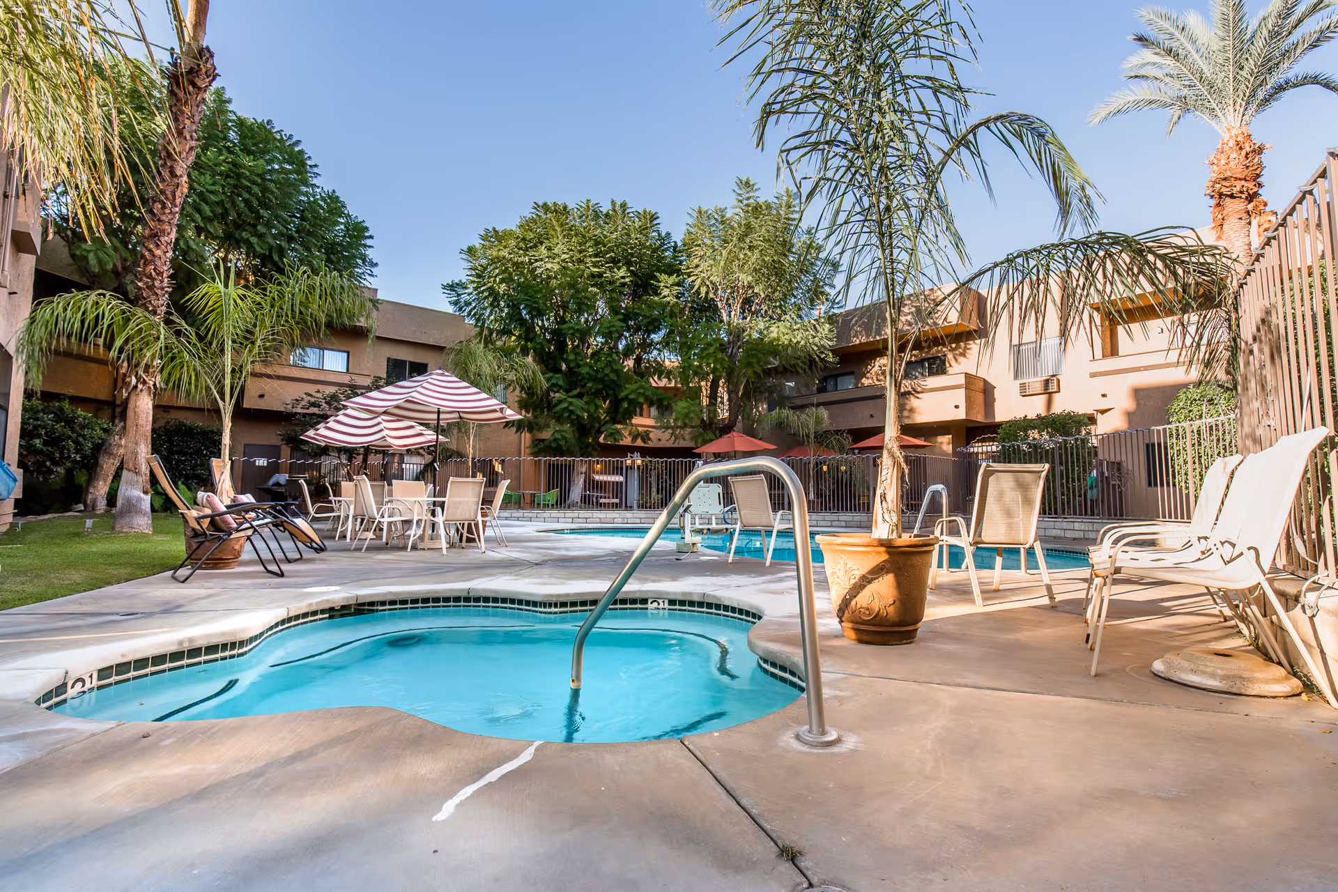 Outdoor pool area at Windsor Court Assisted Living with a small round hot tub in the foreground, surrounded by lounge chairs, tables with umbrellas, palm trees, and a two-story building in the background under a clear blue sky.