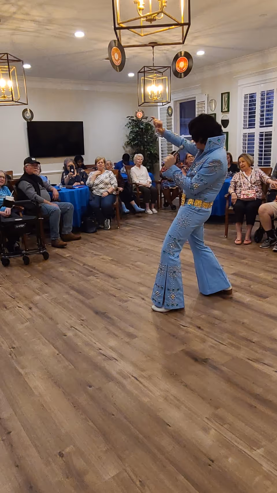 A performer dressed in a blue Elvis Presley-style jumpsuit with gold embellishments is entertaining a seated audience of elderly people in a well-lit room with wooden flooring and modern light fixtures. The audience is seated around tables covered with blue tablecloths, watching the performance attentively.