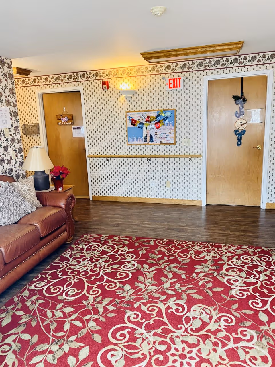 Interior of a senior living facility room with floral wallpaper, a red patterned carpet, a brown leather couch with cushions, a side table with a lamp and a poinsettia plant, two wooden doors decorated with a welcome sign and holiday ornaments, a bulletin board with decorations, and an illuminated exit sign above one door.