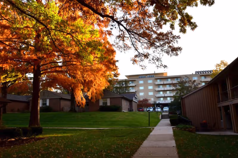 A pathway leading through a grassy area with large trees showing autumn foliage in orange and green. On either side of the path are residential buildings, with a multi-story apartment building visible in the background under a clear sky.