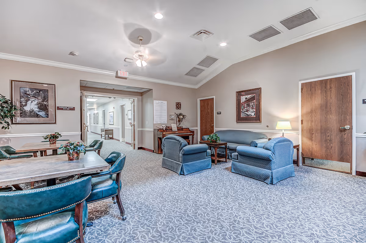 Well-lit senior living common room with upholstered chairs, sofas, tables, and framed artwork on the walls.