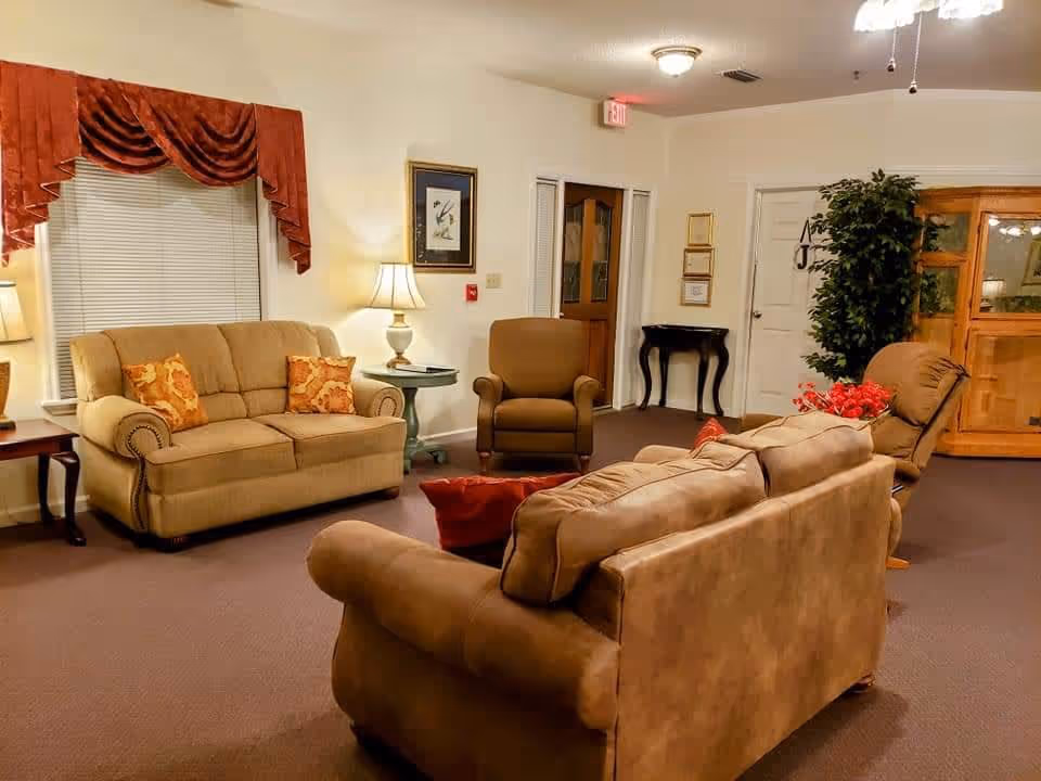 A cozy living room with beige and brown sofas, an armchair, side tables with lamps, a window with red curtains, framed artwork on the wall, a potted plant, and a wooden cabinet. The room has warm lighting and a carpeted floor.