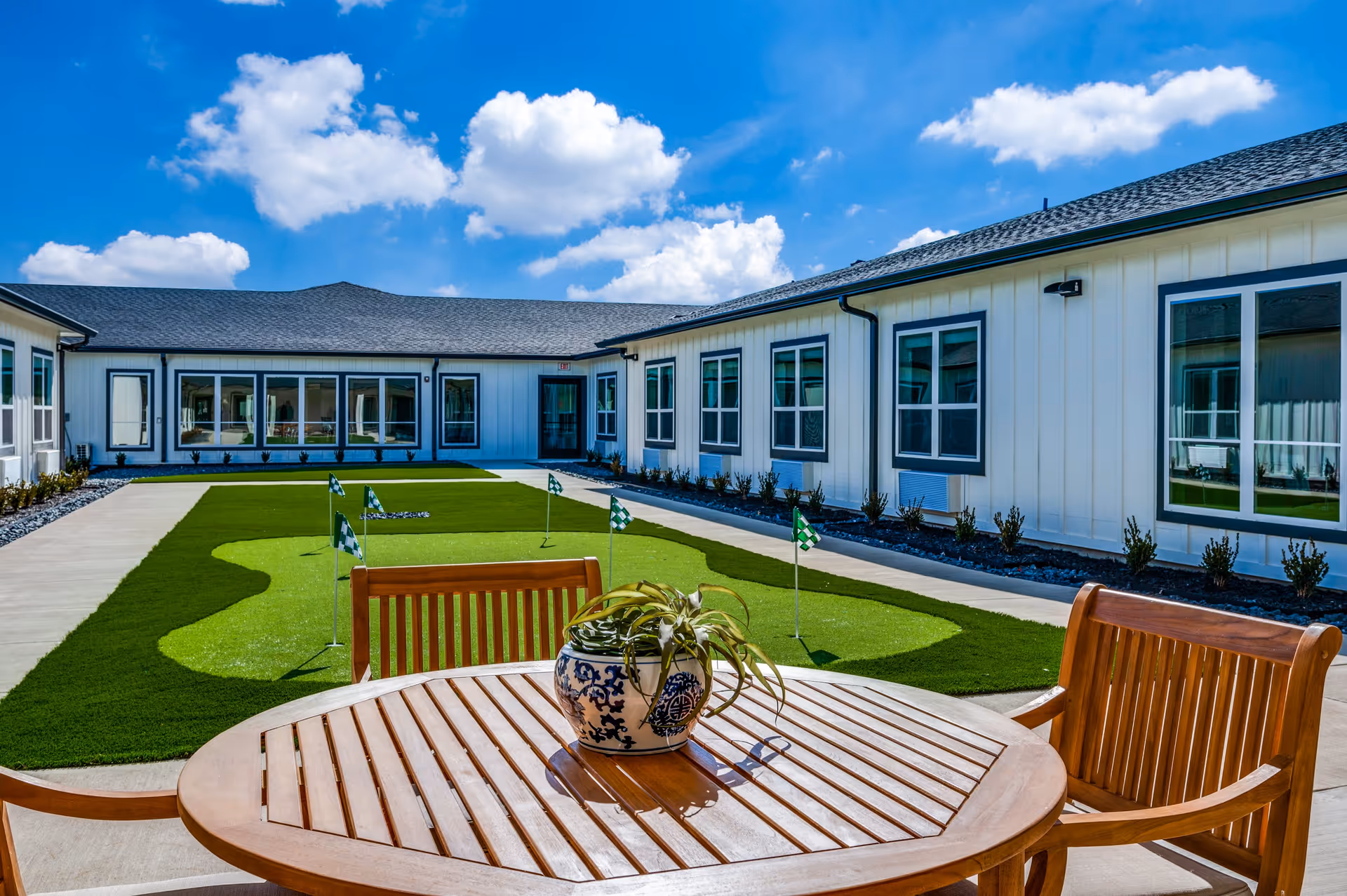 Outdoor courtyard area at The Lodge of Saginaw Health & Wellness featuring a small putting green with several golf flags, surrounded by a concrete walkway and white building with multiple windows. In the foreground, there is a wooden round table with a decorative blue and white ceramic pot containing a plant, and wooden chairs. The sky is bright blue with scattered white clouds.