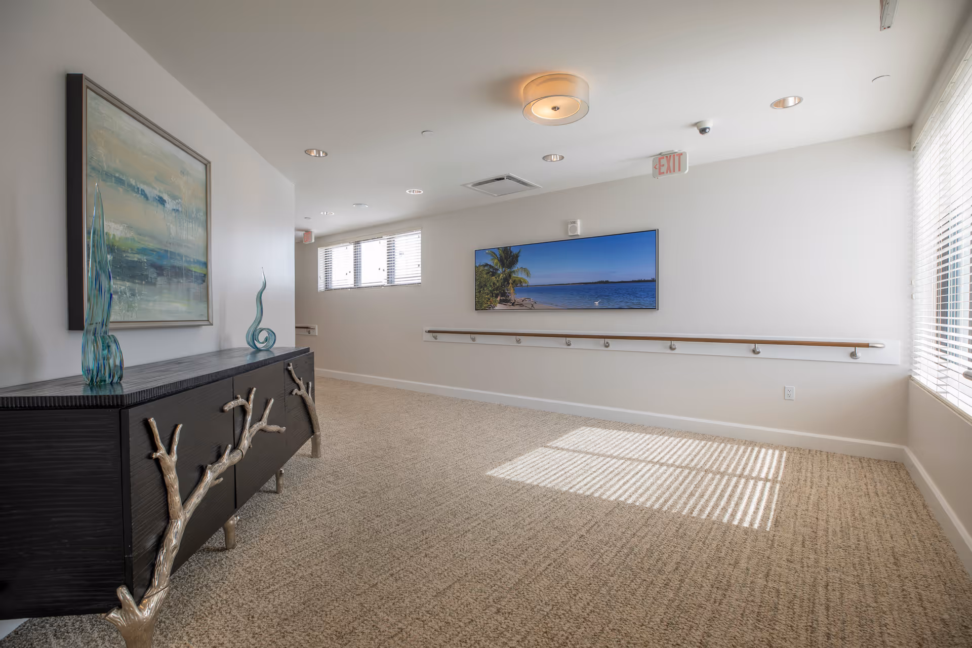Well-lit carpeted hallway with a decorative black console, wall art, a wall-mounted TV showing a beach scene, and a handrail along the wall.