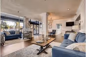 Bright communal living room with blue sofas and armchairs around a wooden coffee table, a rug, large windows, and a wall-mounted TV.