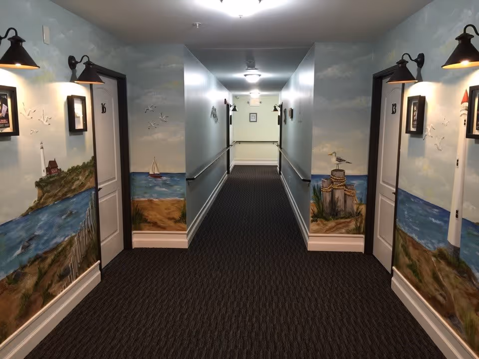 A hallway in a memory care facility with carpeted floor and handrails on both sides. The walls are decorated with murals depicting coastal scenes including a lighthouse, seagulls, a sailboat, and beach landscapes. There are doors numbered 13 and 16 visible on either side of the hallway, each with a wall-mounted light fixture above framed pictures.