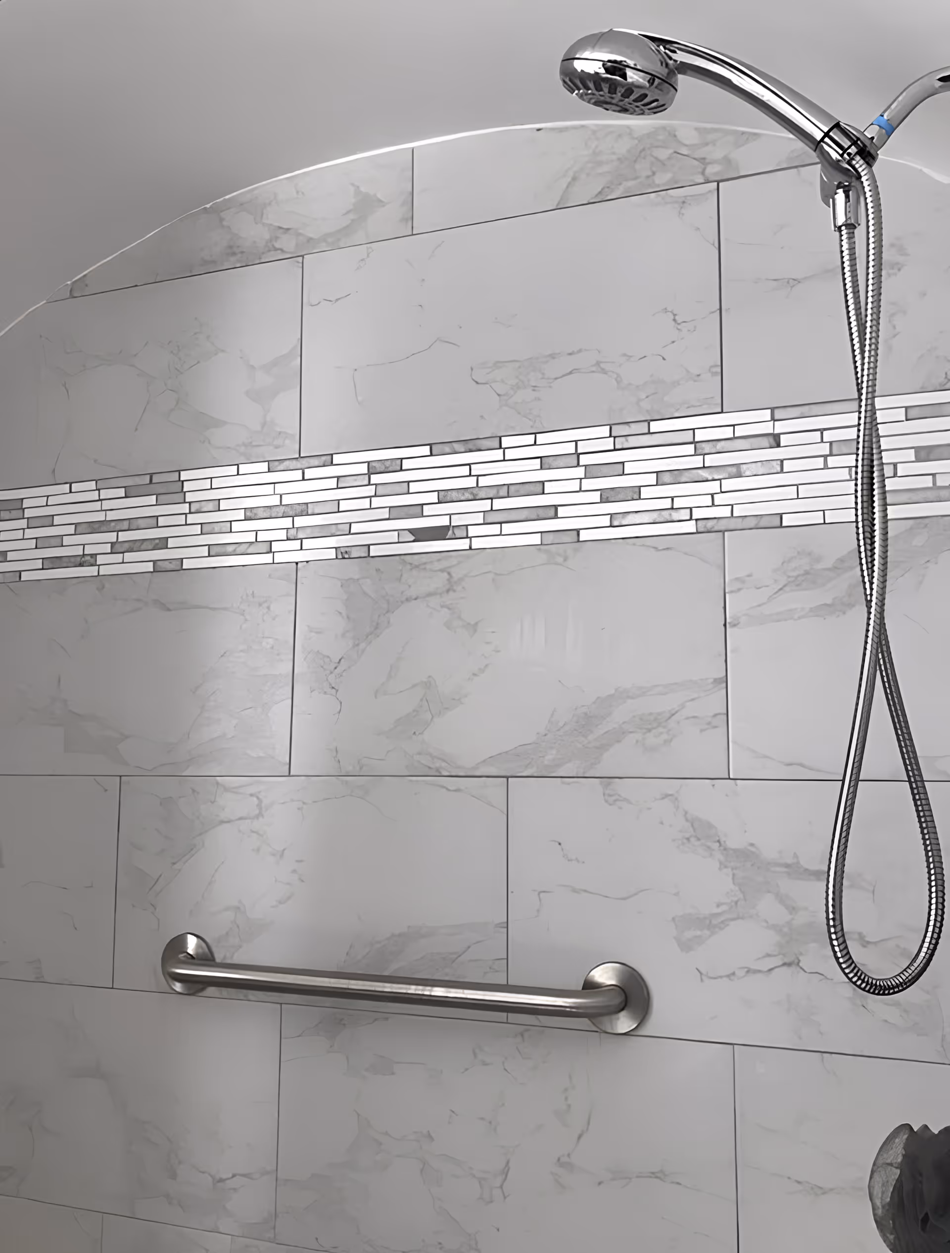Shower area with white and gray marble-patterned tiles, a horizontal strip of small rectangular mosaic tiles, a silver grab bar, and a chrome handheld showerhead with a flexible hose.
