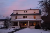 Two-story white house with a covered front porch and a snow-covered yard at dusk.