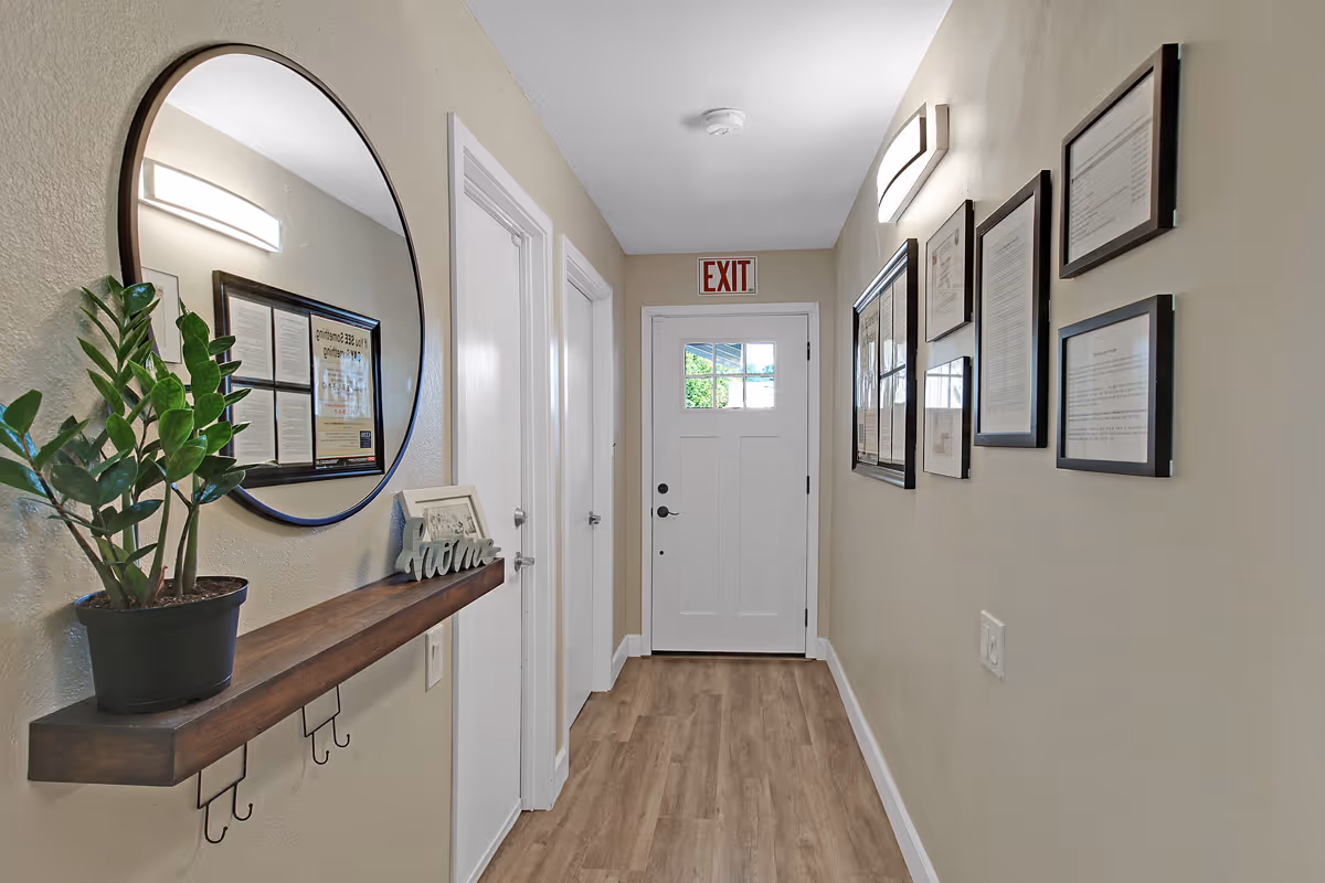 A narrow hallway with light beige walls and wood flooring. On the left wall, there is a round mirror above a wooden shelf holding a potted plant and a decorative 'Home' sign. Two closed white doors are visible on the left side. The right wall features several framed documents or pictures. At the end of the hallway, there is a white door with a window and an illuminated exit sign above it.